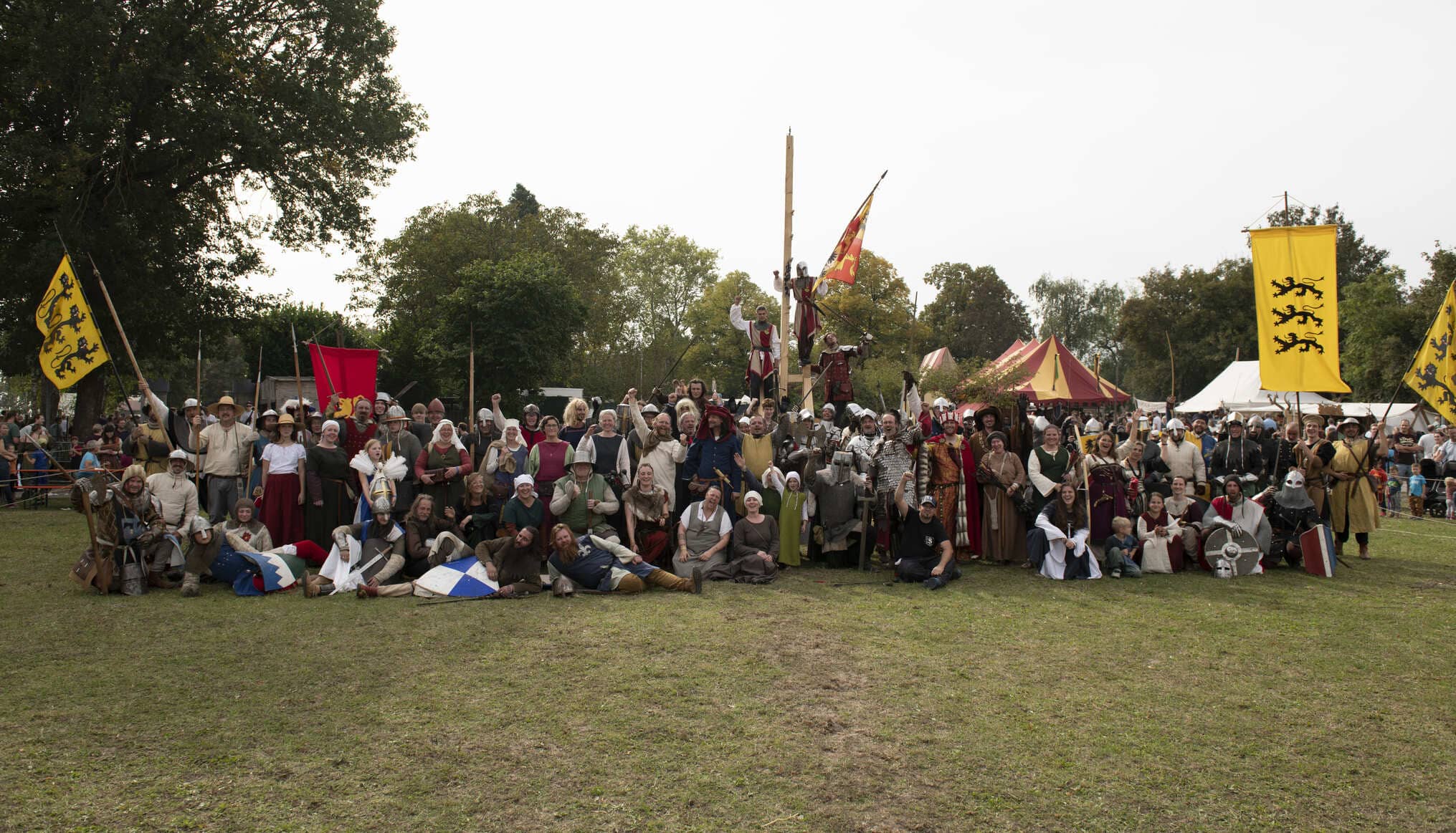 Große Gruppe von Teilnehmenden in mittelalterlicher Kleidung und Rüstung auf einer Wiese beim Burg Stettenfels Mittelalterfest, umgeben von Bannern und Zelten.